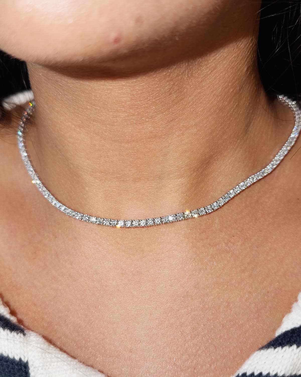 A white gold tennis necklace of four prong set lab grown diamonds, worn close at the base of the neck